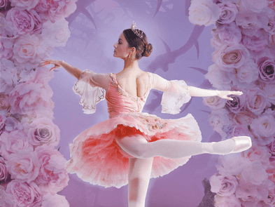 A ballet dancer in pink ballet outfit and white ballet shoes raises one leg mid twirl in front of a background of pink blossom