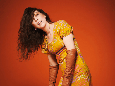 Bridget Christie wearing a yellow dress and leaning to the side in front of a brown background. 