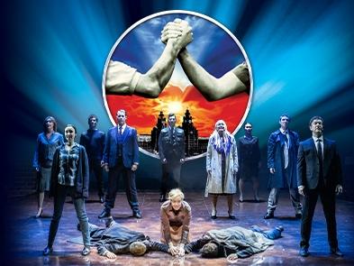 Two actors lie face down on the floor with another actor kneeling down between the two of them with other cast members standing behind them, the blood brothers logo, two hands clenching each other, is above the scene