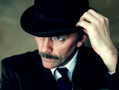 Man with moustache holding on to bowler hat on head, looking down with serious expression