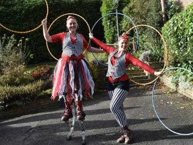 Two women are wearing red, blue and white stripy circus outfits and holding big hoops. One of them is standing on stilts. 