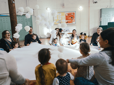 kids and adults sitting around a white sheet holding onto it. 