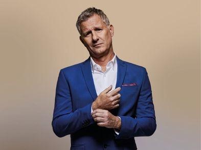 Adam Hills - a white male with short dark blonde hair, is wearing a blue suit and white shirt with arms folded holding his cuff, looking perplexed. 