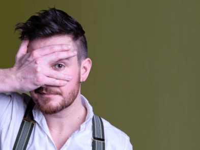 A man with black hair peering through his fingers against a green background