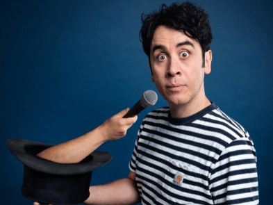 Pete Firman wearing a black and white t-shirt holding a top hat with a hand and microphone coming out of it.